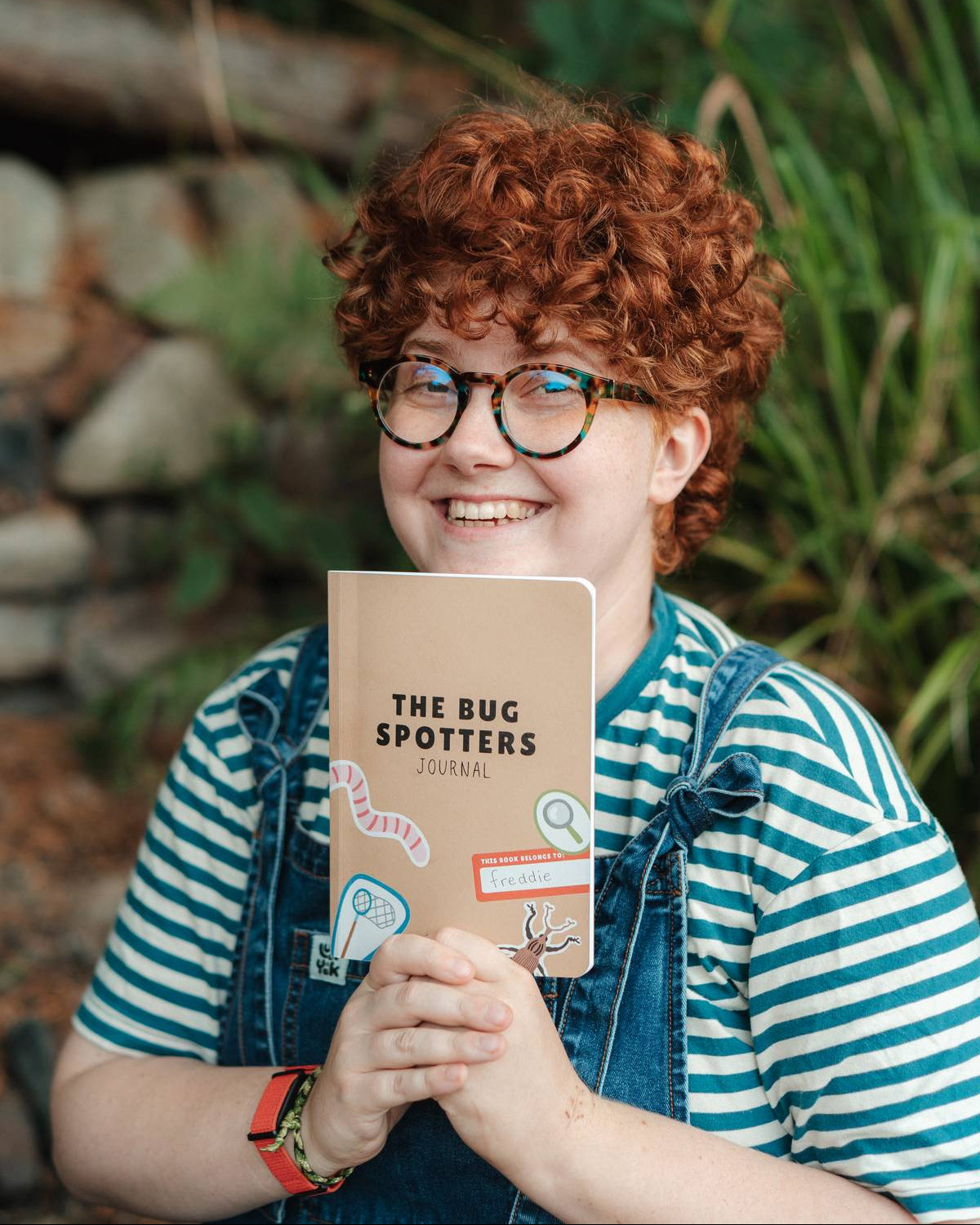 A smiling person with curly red hair and glasses holds a notebook in front of their face. The person is wearing a blue and white striped shirt under denim overalls, with a blurred outdoor background of green plants and rocks.