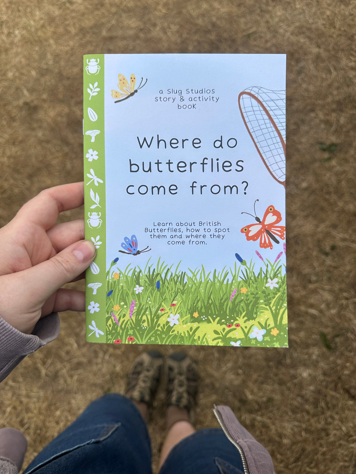 A hand holds a children's activity book titled 'Where do butterflies come from?' outdoors on a grassy ground. The book cover features a light blue top half with black text and a green bottom half with grass, flowers, and colorful butterflies.