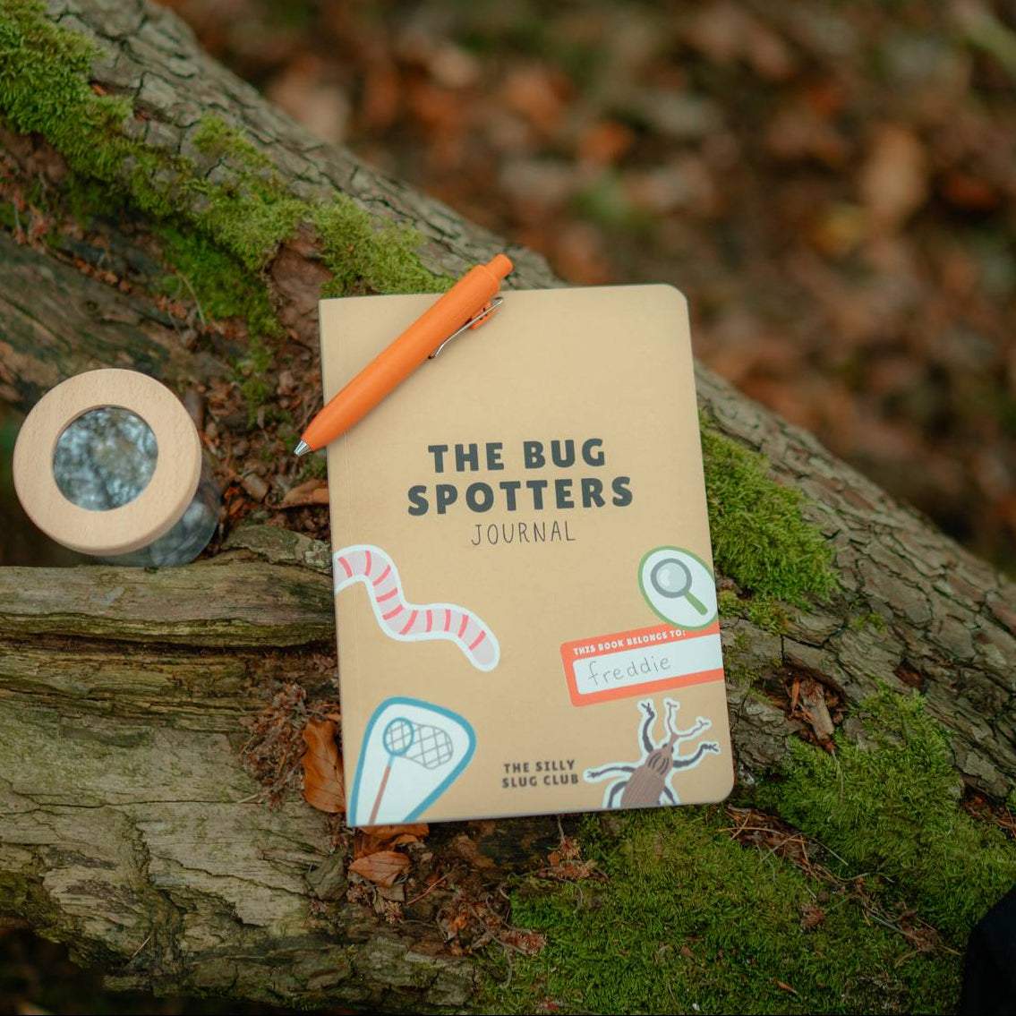 A beige softcover journal titled 'THE BUG SPOTTERS JOURNAL' lies on a mossy tree branch outdoors, with an orange pen resting on top and a small round bug observation container nearby.