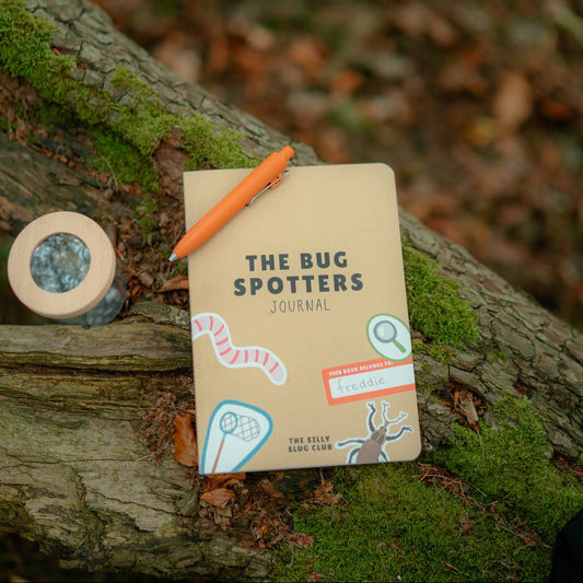 A beige softcover journal titled 'THE BUG SPOTTERS JOURNAL' lies on a mossy tree branch outdoors, with an orange pen resting on top and a small round bug observation container nearby.
