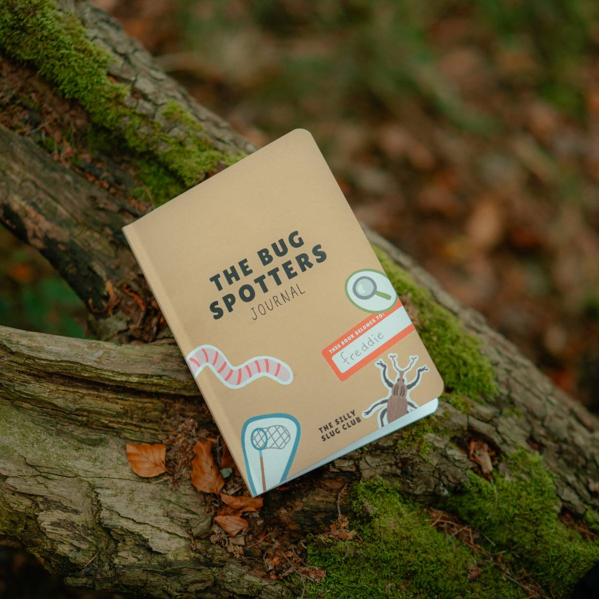 A book titled 'The Bug Spotters Journal' resting on a mossy tree branch in a forest setting.
